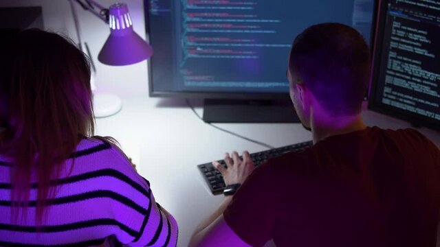 Two smart young programmers are coding on computer in process of discussion. Back view of man and woman sitting in front of monitor with java script and typing. Theme of website design and cyberspace.