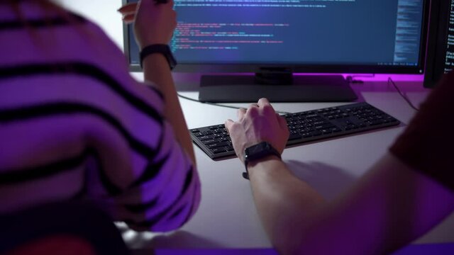 Team work of two programmers who are sitting in front of computer screen and typing javaskript in closeup. Back view of colleagues who are discussing the job points and writing new lines of html code.