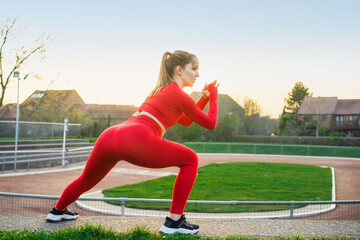 Back view Young fitness woman in red sport wear doing side lunge, squats during outdoors workout on...
