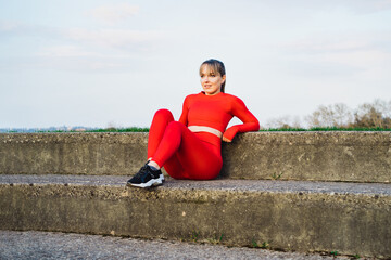 Portrait of a smiling confident fitness woman in red sport wear resting after work out or running outdoors on sunset. Jogging in the stadium. Active lifestyle and health care. Copy space. © okrasiuk