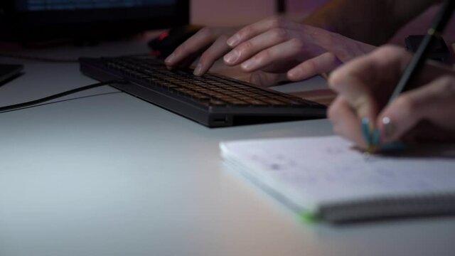 A man and a woman are sitting in office in front of computer and developing new project in macro. A lady writes notes and a male person types on keyboard. They talk and gesticulate. Team work concept