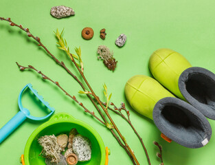 Children's rubber boots, a plastic bucket with finds from the forest, park and branches with buds and green leaves. On a green background, overhead