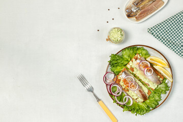 Two rye crisp bread with sardines and cream cheese and herbs, on a gray plate with green lettuce leaves. On a light gray background, horizontally with space