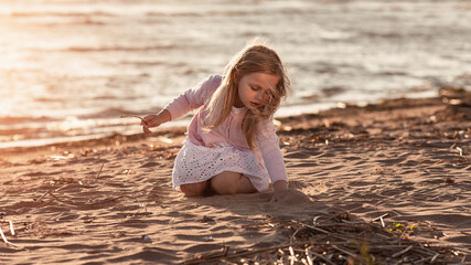 Portrait of little cute blond girl drawing with a stick on the sand at the sunset beach
