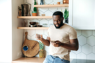 Focused young African American stylish bearded man standing at kitchen, holding a glass of pure...