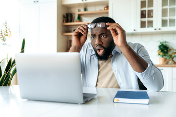 Shocked African American bearded businessman, manager or broker, sits at his desk, looks in...