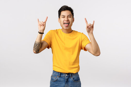 Excited Attractive Young Asian Guy Having Fun, Dancing And Enjoying Stunning Concert, Smiling Showing Tongue, Make Rock-n-roll, Heavy Metal Signs, Adore Amazing Song, White Background