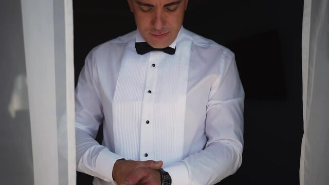 An elegant manly caucasian man stands in formal wear in a white shirt and classic trousers and puts on an accessory - a stylish watch to see the time. Fashionable, expensively dressed businessman