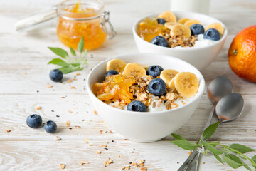 bowls of granola and yogurt with orange jam, bananas and blueberries on a light table