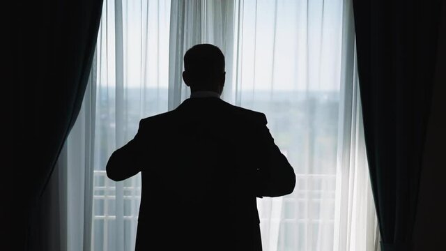 An elegant man stands by the window with curtains a day and puts on a jacket in preparation for an important event. Successful male businessman wearing a suit. Silhouette of a man behind
