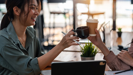 Happy working Asian woman hold coffee cups and cheers at café.