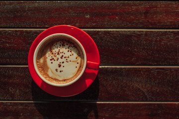 Red cup of hot latte coffee on wooden table at sunrise.