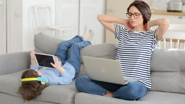 Family, quarantine, distance job concept. Mother freelancer resting after work from home office on laptop during lockdown, sitting on couch, little son playing on digital tablet and wear headphones. 
