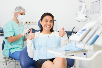 Happy cute latin woman sitting in dental chair after teeth cure giving thumb up