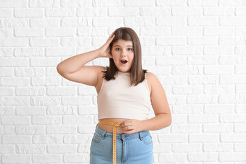 Shocked overweight girl measuring her waist on white brick background