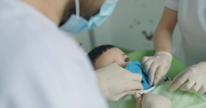Close-up a little boy that laying in the dental chair with an open mouth and wearing a rubber dam on his teeth while the dentist is fixating the rubber dam by using white plastic detail.