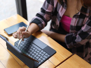 Female teenager using digital tablet with stylus and keyboard on wooden table in cafe