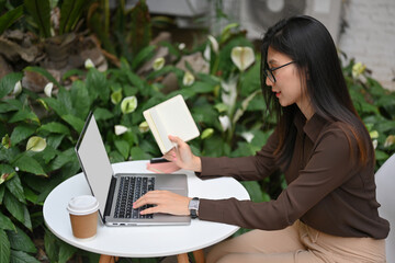 Female holding schedule book and working with laptop on round table in garden at home