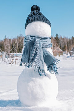 A snowman in a blue hat and scarf is standing in the field. An iceman on a winter forest background. Sunny day, freezing cold, close up.