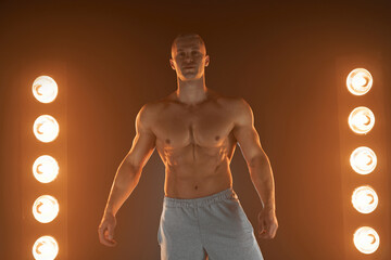 Active lifestyle. Professional bodybuilder showing perfect muscular body and smiling at camera, lamps illumination on background