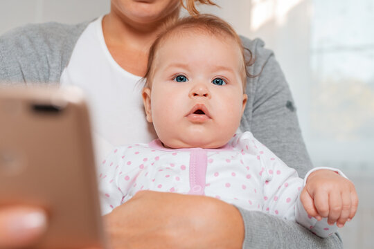Close-up portrait of a baby sitting in the arms of a mother who uses a smartphone. Bottom view