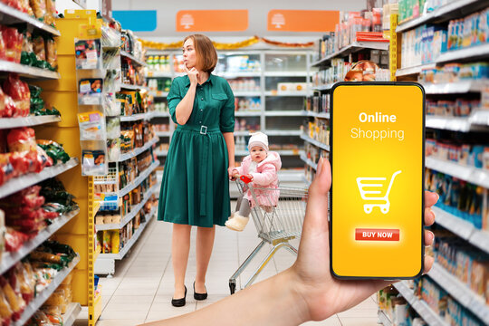 Shopping. A Young Woman Chooses Products, Holding A Grocery Cart With A Baby Sitting In It. View Among The Supermarket Shelves. The Concept Of Online Shopping And Consumerism