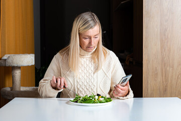 middle aged blonde woman  using smartphone while eating salad in the kitchen