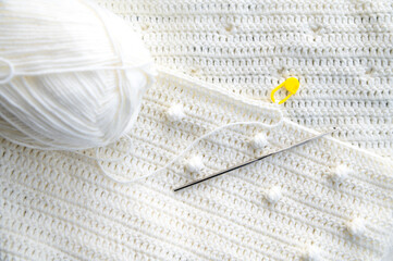 A fragment of a crocheted plaid is folded several times. The loop of the unfinished knitted row is fixed with a yellow holder. On the canvas is a metal hook for knitting and a ball of thread