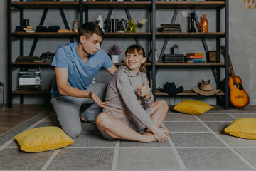 Couple doing Yoga at home in pajamas near bookshelf. Young man and woman doing sports at home.