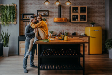 Man hugging woma in the kitchen. Woman in yellow sweater sitting on the table. Happy couple in kitchen.