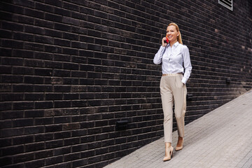 Full length of attractive smiling fashionable woman walking outdoors and talking on the phone.