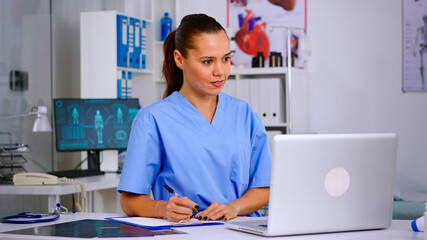 Doctor assistant in uniform working on laptop in modern hospital office. Therapist writing list of consulted patients online, making research, noting, taking useful information on clipboard