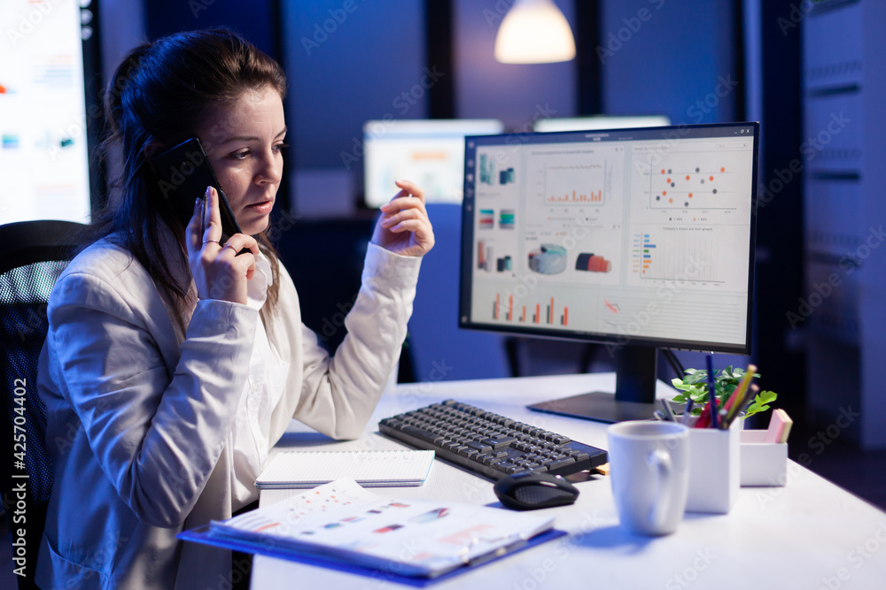 Poster businesswoman speaking at smartphone with her teamate while working at computer with graphic financi