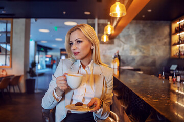 Bossy and beautiful woman in business clothes sitting on a break in a cafe of a hotel and holding a cup of coffee. Break between two meetings. Business lifestyle, hotel stay over, coffee break