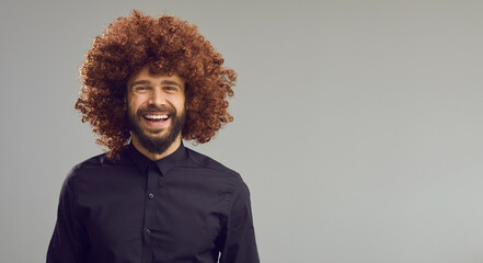 Obraz premium Happy young guy with thick curly brown hair on blank advertising space for text. Funny laughing man in Afro wig standing on light grey copy space banner background