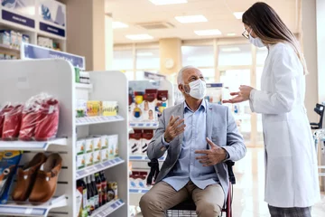Foto op Canvas A man in a wheelchair talks to a female pharmacist in white uniform at the pharmacy. Discussion of medical therapy and sale of drugs. They both wear a protective face mask due to the corona virus © dusanpetkovic1