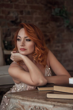 Pretty slender woman with beautiful red hair. Female in stage image at table with books and candles. Emotions girl in dress sitting in interior old textured room. Concept of learning and knowledge