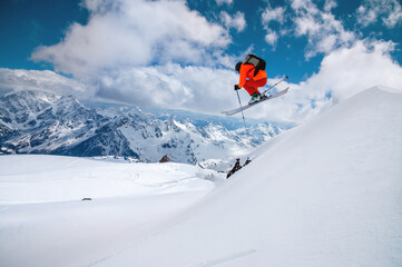 A freerider skier in an orange suit with a backpack froze in a jump flight over high snow-capped...
