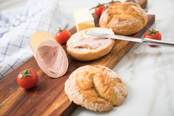 Feine Kalbsleberwurst auf Weizen Rosen Brötchen dazu Tomaten, Butter und Messer mit Holzbrett, Geschirrtuch und Marmor Hintergrund als Brotzeit Vesper