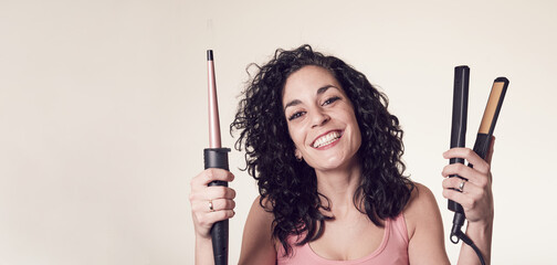 Smiling curly-haired young woman can't decide between using his curling iron or his hair straightener. care and beauty concept copy space.