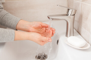 Women's hands close-up under running water in the bathroom above the sink. There's a piece of soap nearby