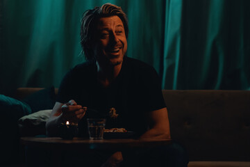 Smiling blond man in black t-shirt sitting on a sofa behind a coffee table at dinner watching the television.