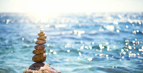 Obraz premium A pyramid of pebbles on a large stone against the background of the sea and the sun's glare on the water. Tour, tourism, vacation at the resort, swimming. Travel background, sunscreen. Copy space