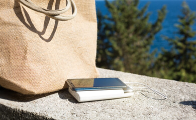 Power bank charges a smartphone on the sea front near the beach bag mock up. A portable travel...