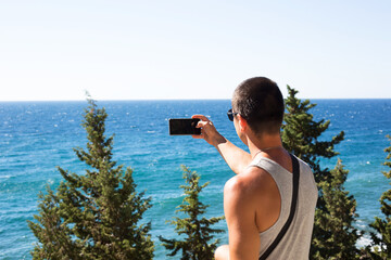 Male tourist takes photos on a smartphone of beautiful view of sea in a mountainous area from the top among coniferous trees. Video communication, using the phone when traveling. SIM card in roaming