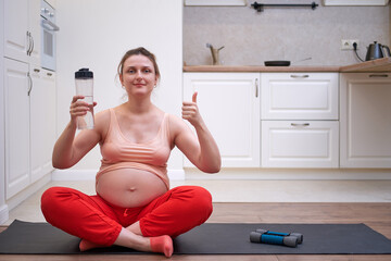 Fitness during pregnancy and the benefits of clean water. Pregnant woman at sports training at home shows the thumbs up with a bottle of water in her hands