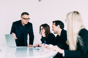 Confident team leader giving presentation at corporate meeting in modern boardroom. Business people sitting at table, listening to coach mentor explaining strategy