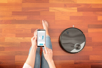 Top view of faceless middle section of young woman using smart phone to control an automatic vacuum...