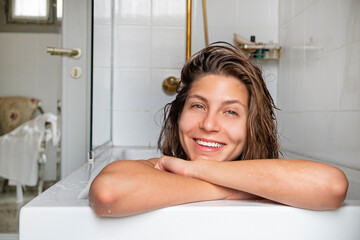 Obraz premium Close up portrait of a beautiful smiling young woman relaxing in a bathtub. Happy brunette female chilling in a bath of hotel room. Copy space, background.