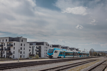 Modern white and blue passenger train on a station in front of a modern housing or residental district. City blocks and train.